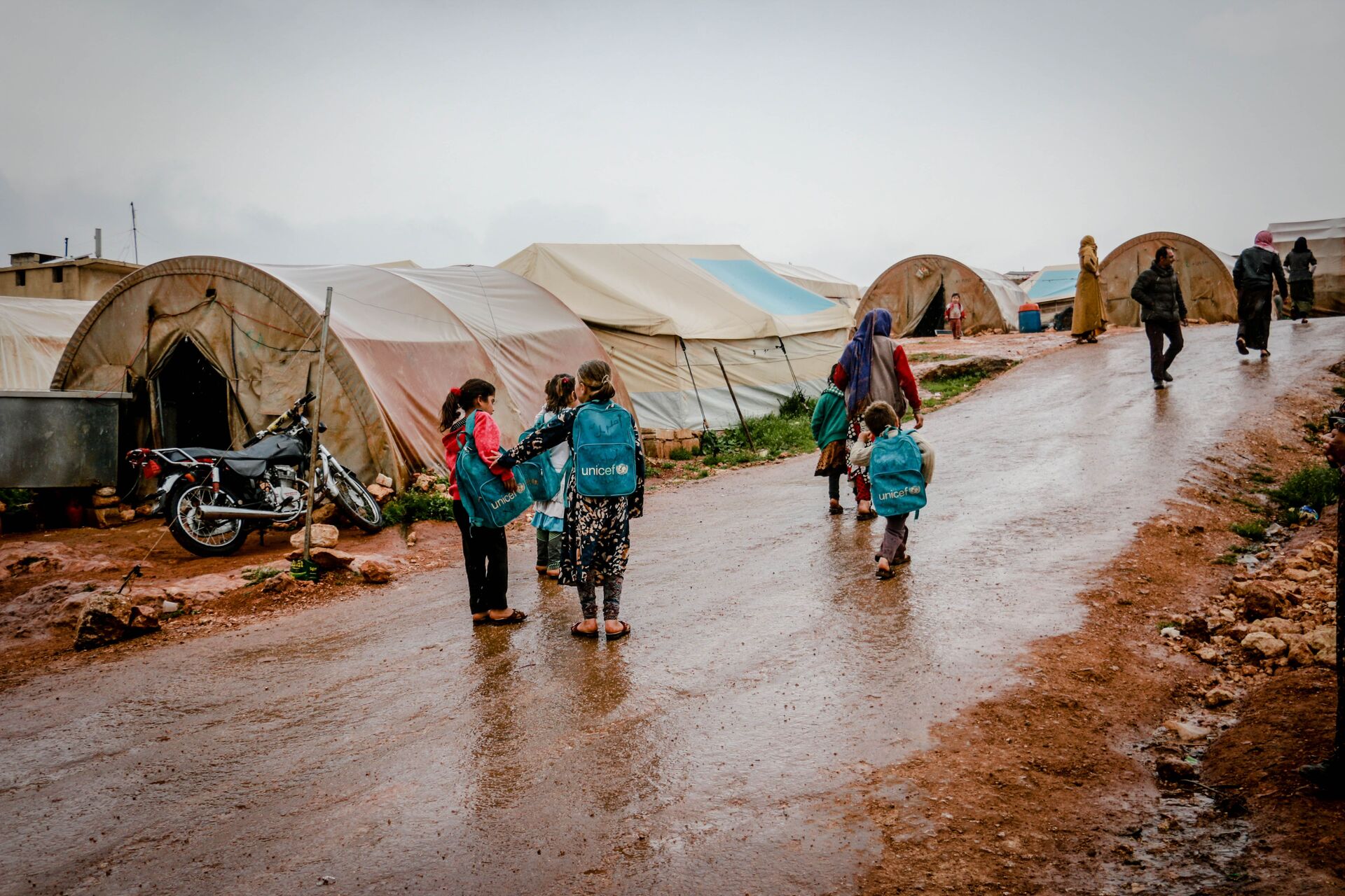 Children walking along a muddy path between tent structures in a community camp